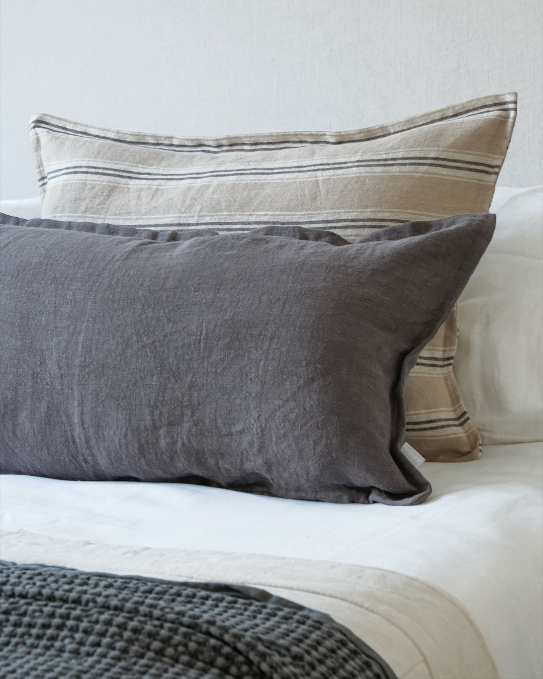 Funda de cojín Kankla de lino gris oscuro con borde de pestaña, sobre una cama con cojines decorativos de rayas.