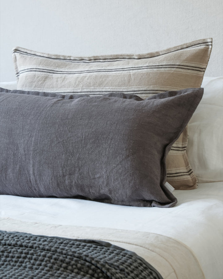Funda de cojín Kankla de lino gris oscuro con borde de pestaña, sobre una cama con cojines decorativos de rayas.