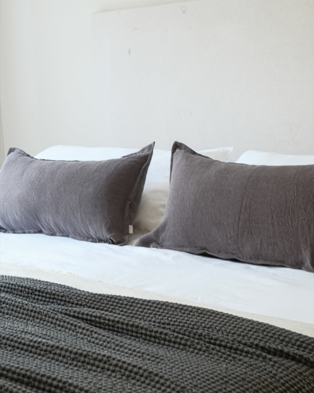 Funda de cojín Kankla de lino gris oscuro con pestaña decorativa sobre una cama blanca