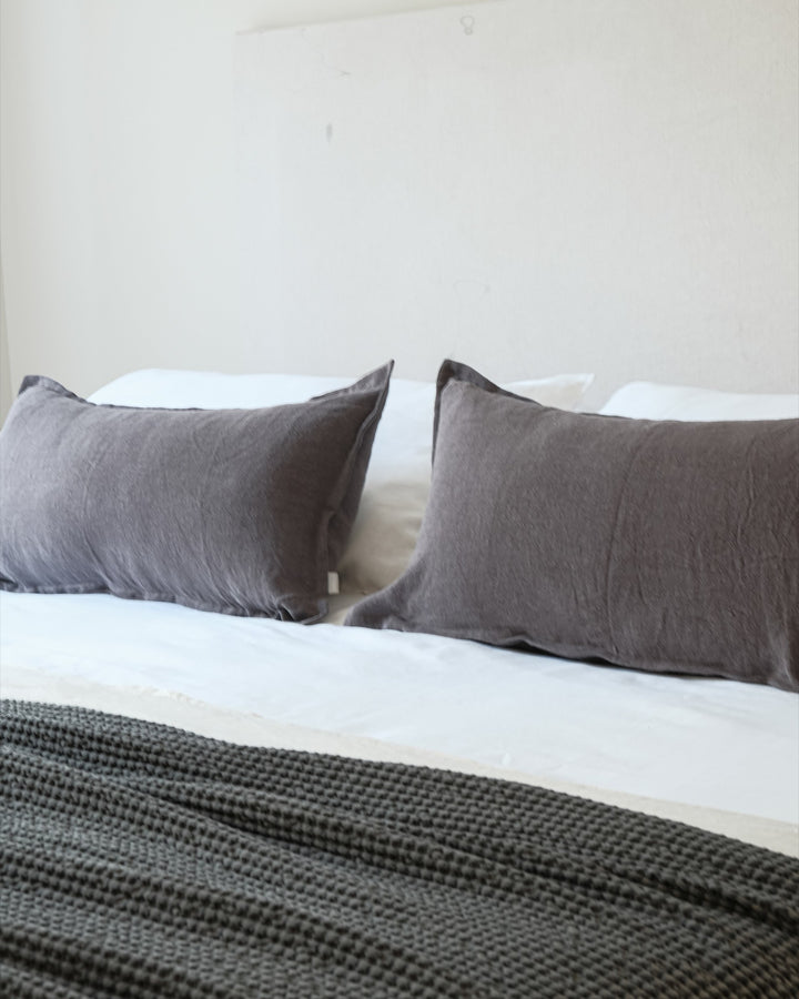 Funda de cojín Kankla de lino gris oscuro con pestaña decorativa sobre una cama blanca