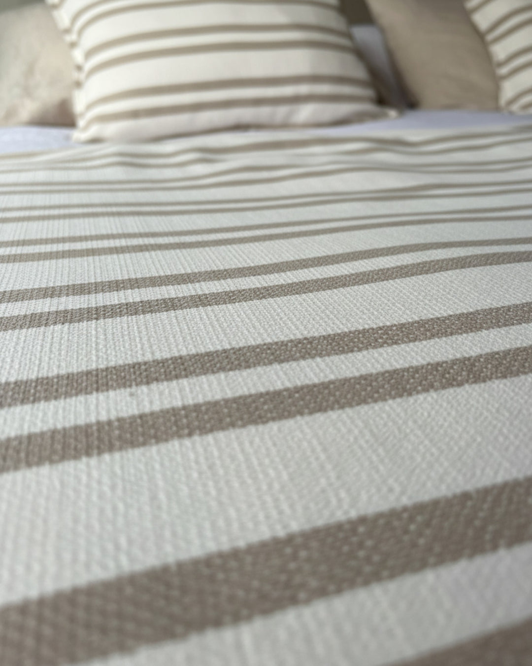 Piecera Kankla Mónaco reversible de algodón con diseño de líneas beige y lino neutro sobre cama con cojines