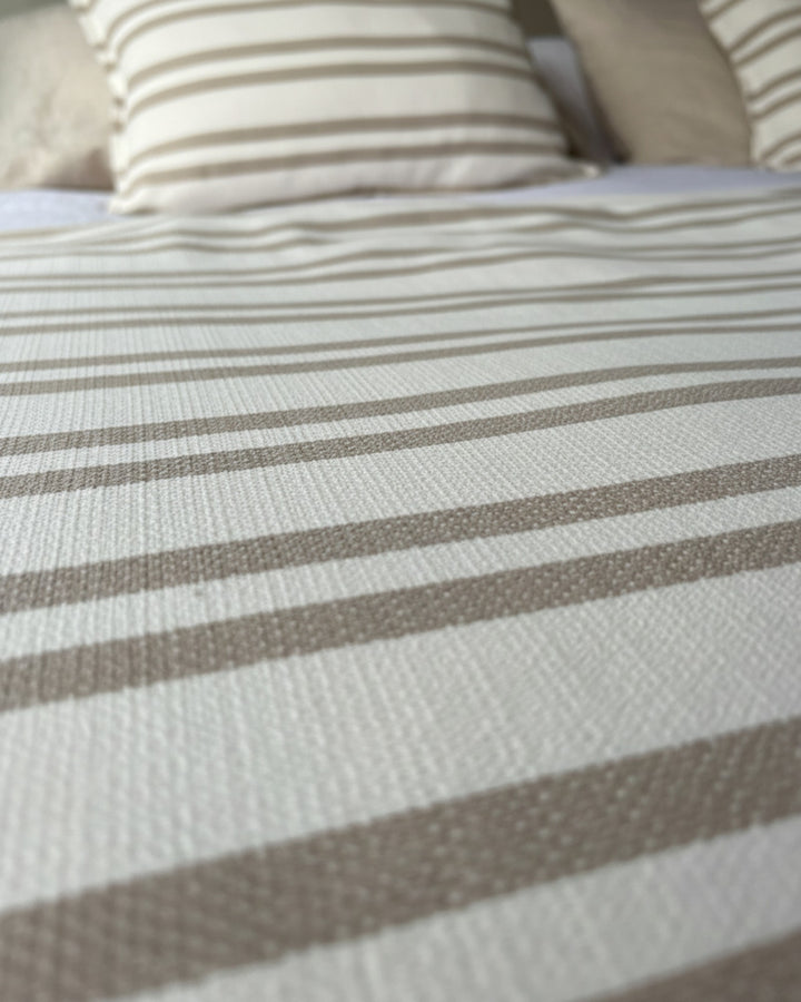Piecera Kankla Mónaco reversible de algodón con diseño de líneas beige y lino neutro sobre cama con cojines